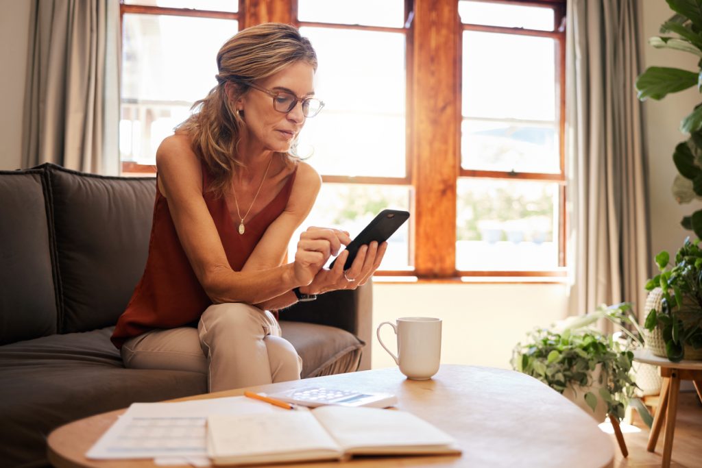 Budget, smartphone and senior woman with finance paper, mobile app or website information for retirement planning, tax or asset management. Elderly person using phone for financial tech on home sofa